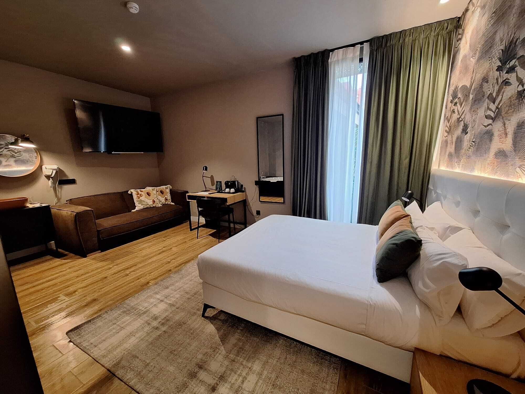 Habitación de hotel con cama, escritorio, silla, televisor y ventana con cortinas.