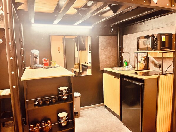 A compact kitchen with a microwave, sink, and a coffee machine. There is a shelf with mugs and a trash bin. - Casa fresco 1 (Nîmes)