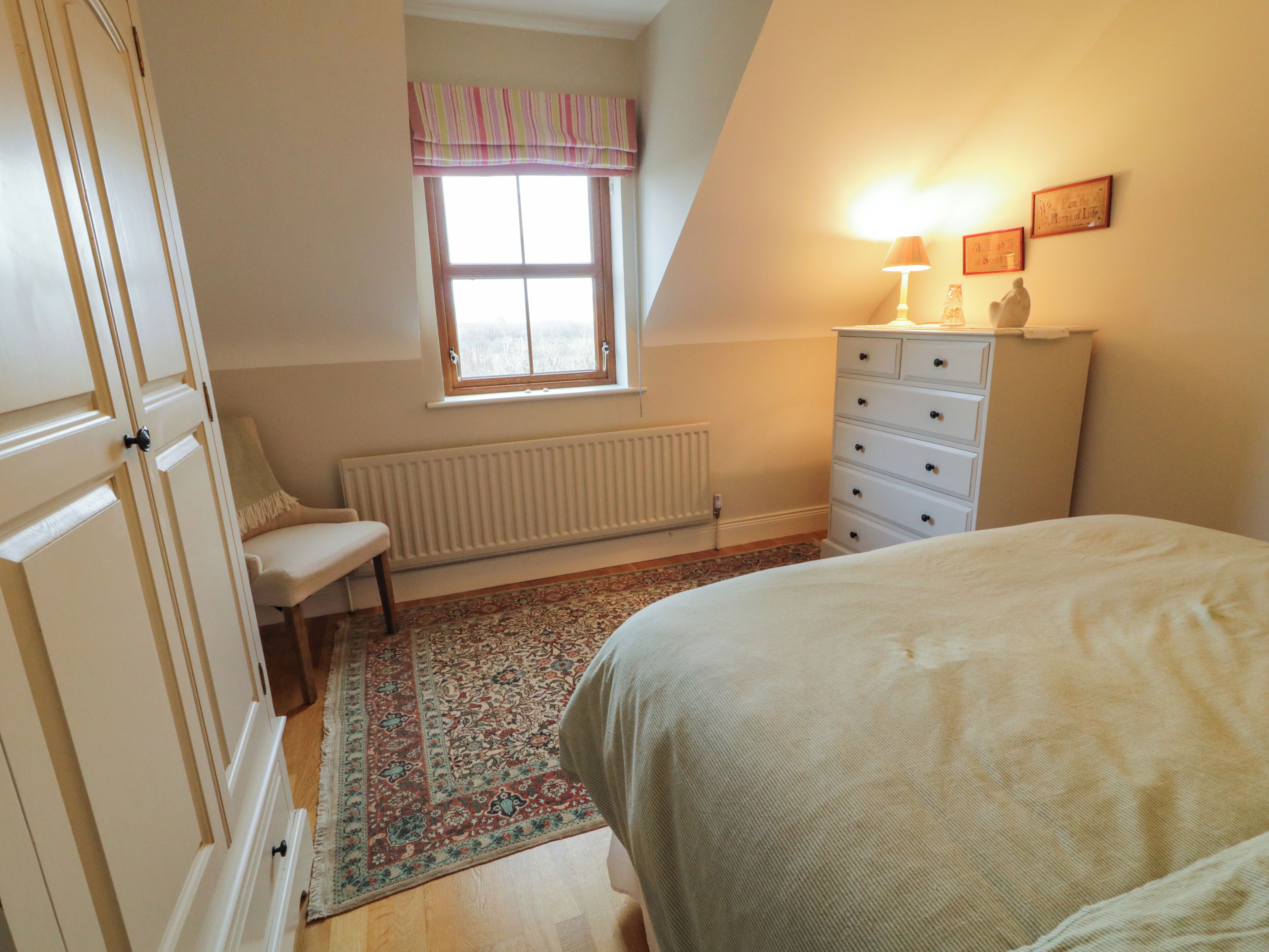 Une chambre à coucher comprenant un lit, une commode, une chaise, une fenêtre avec des rideaux et un radiateur.