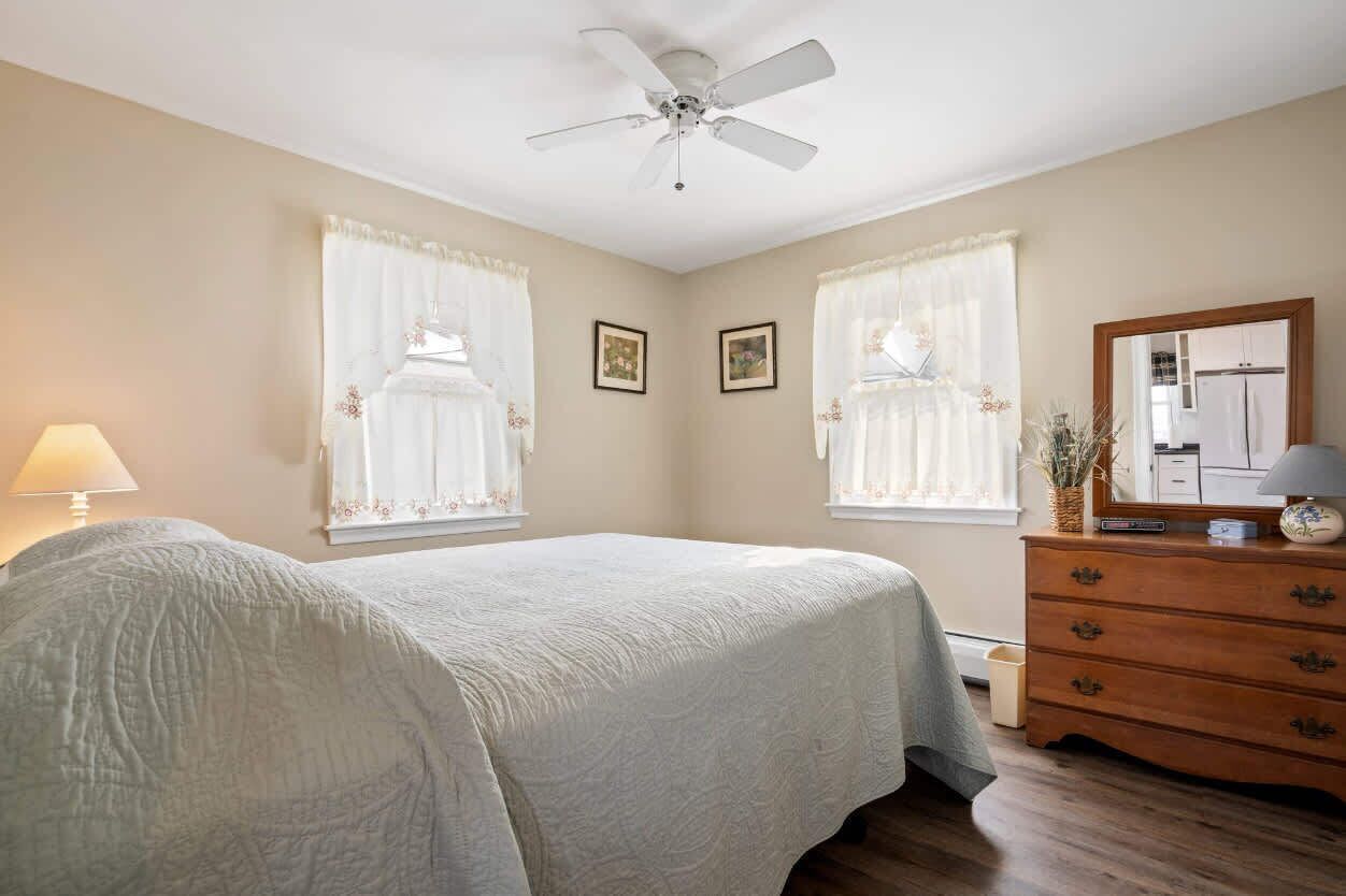 A bedroom with a bed, a dresser, a ceiling fan, and two windows with curtains.