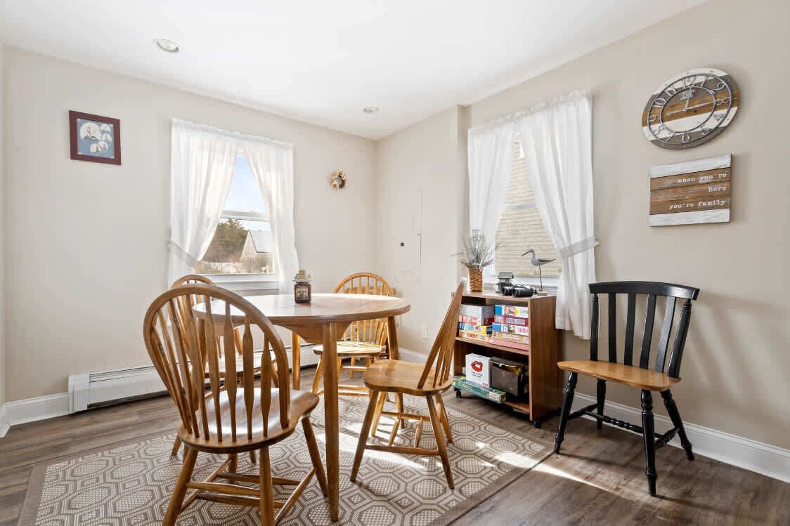 A dining area with a round table, wooden chairs, and a black chair. There are windows with white curtains, a wall clock, and a sign with a message.