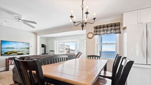 A dining area with a wooden table and chairs, a chandelier, and a view of the ocean through large windows. - Wright by the Sea For 10! (York Beach)