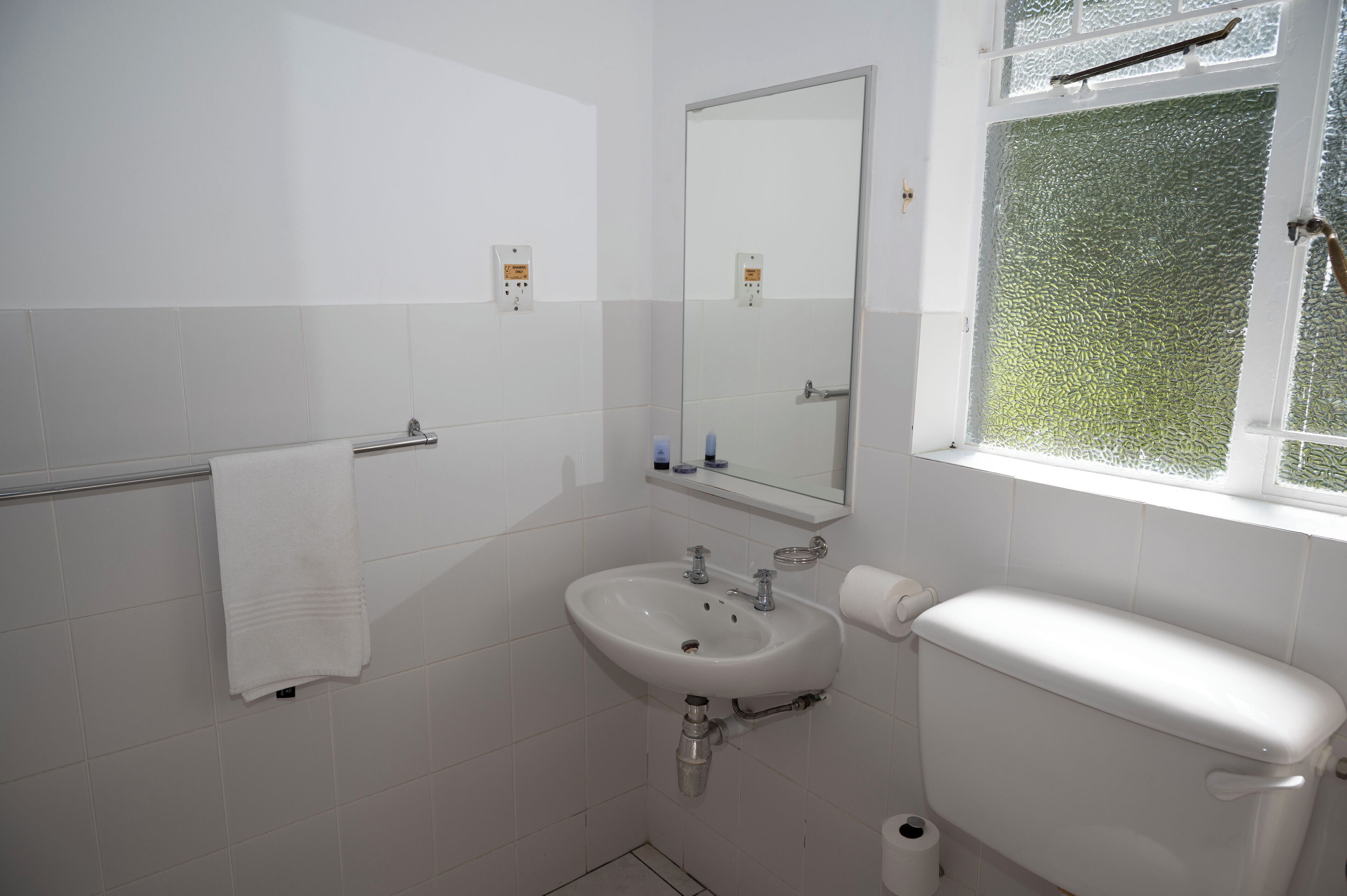 A bathroom with a white sink, mirror, and toilet, with a towel rack and a window with frosted glass.