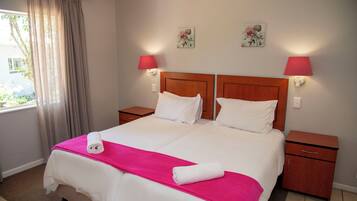 Una cama doble con sábanas blancas y una manta rosa, dos mesitas de noche, un ventanal con cortinas y dos lámparas de pared.