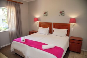 Un llit doble amb llençols blancs i una manta rosa, dues taules de nit, una finestra amb cortines i dues làmpades fixades a la paret.