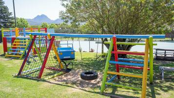 A playground with colorful equipment, including swings and a climbing structure, set against a backdrop of trees and a distant mountain.