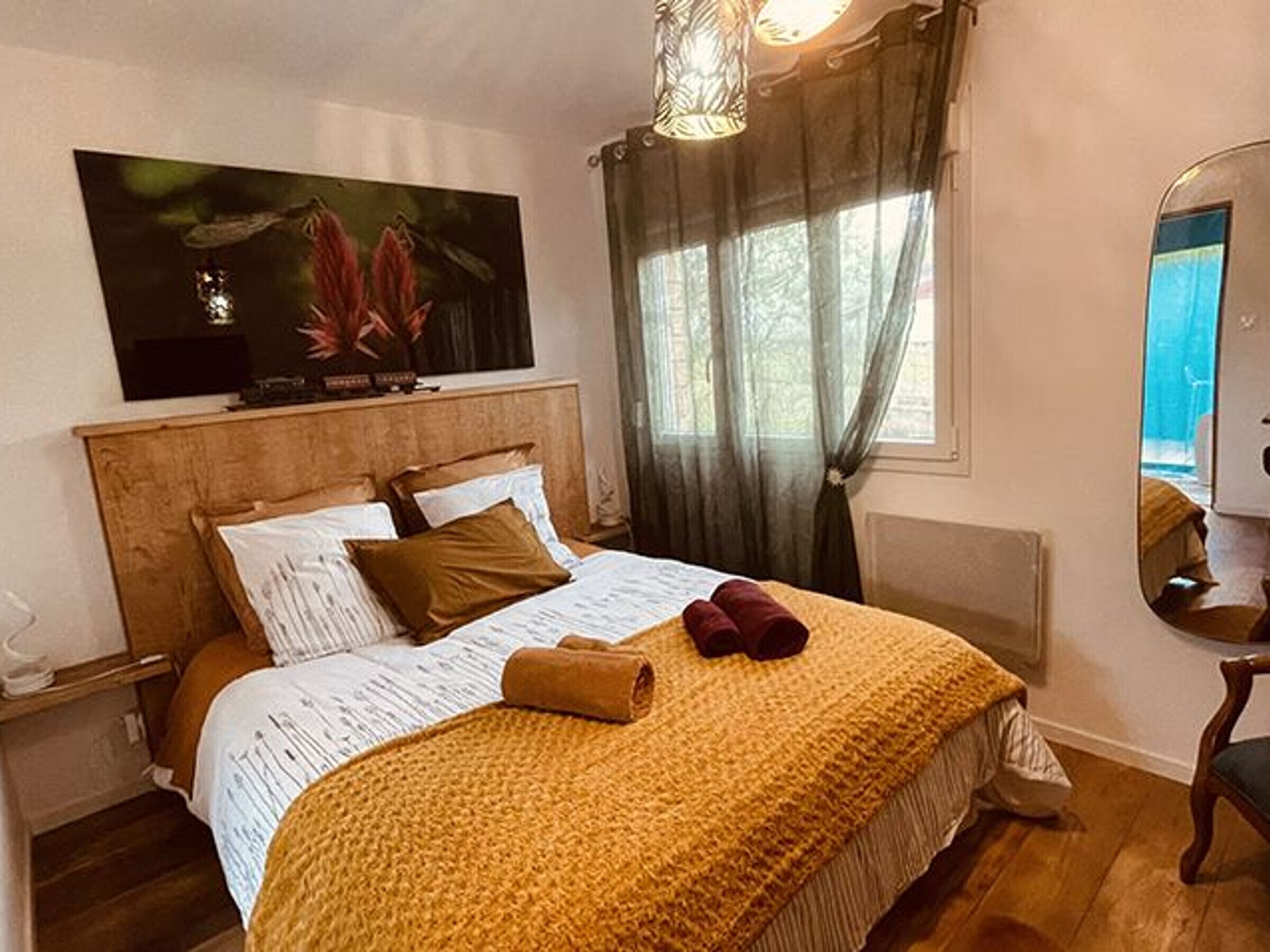 Un dormitorio con cabecera de madera, cama con ropa de cama blanca y marrón, una manta amarilla, un cuadro sobre la cama y una ventana con cortinas.