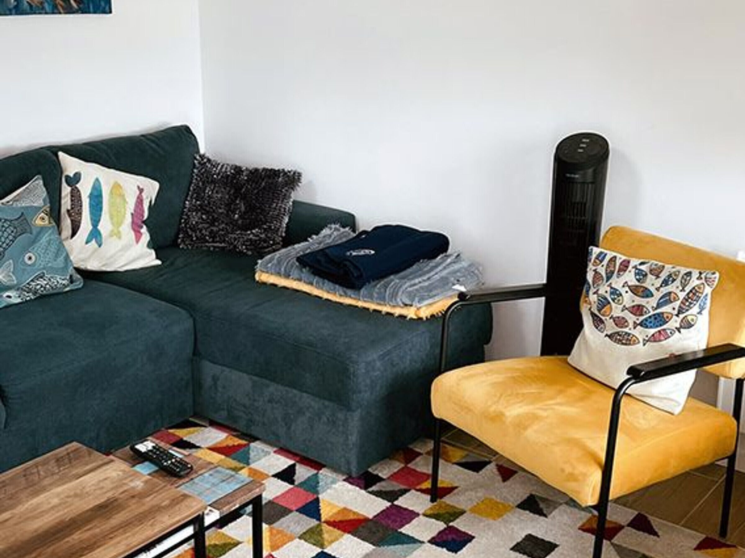 Uma sala de estar com um sofá verde escuro, uma poltrona amarela, uma mesa de centro e um tapete geométrico colorido.