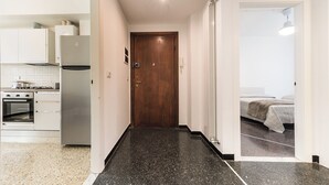 Un couloir étroit, avec une porte en bois, mène à une chambre à coucher où se trouvent un lit et une fenêtre. Une cuisine aux armoires et aux appareils électroménagers blancs est visible sur la gauche.