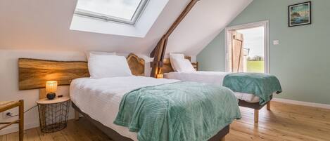 Une chambre avec deux lits, une lucarne, des poutres en bois, une petite table de chevet avec une lampe et une vue sur l’extérieur.