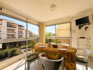 Un comedor con una mesa preparada para cuatro, sillas, un televisor en la pared y vistas a edificios y zonas verdes al exterior.