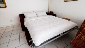 A single-bed room with a metal frame, white bedding, a red chair, a floor lamp, and three floral wall art pieces.