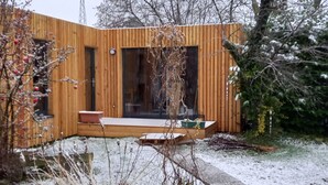 Une cabane moderne en bois, dotée d’une grande porte vitrée, d’un petit porche et d’un jardin recouvert de neige.