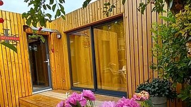Un bâtiment en bois avec une porte en verre, un petit porche et des fleurs roses au premier plan.