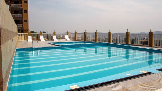A rooftop swimming pool with clear blue water, surrounded by a metal fence and white lounge chairs. - Kololo Business Suite-Pool, Gym, fast internet/ Work, Relax & Unwind (Kampala)