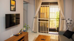 A living room with a flat-screen TV, a wooden coffee table, a sofa, a sliding glass door with curtains, a fan, and a potted plant. - Kololo Business Suite-Pool, Gym, fast internet/ Work, Relax & Unwind (Kampala)