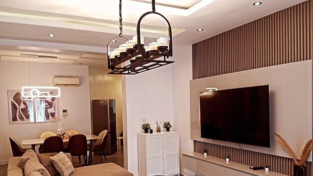 A modern living room with a sectional sofa, a flat-screen TV mounted on the wall, and a dining area with a table and chairs. - Gemluxe Homes (Lagos)