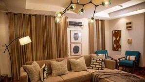 A cozy living room with a beige sofa, a coffee table, and a blue armchair. There is a floor lamp and a ceiling light fixture. - Gemluxe Homes (Lagos)