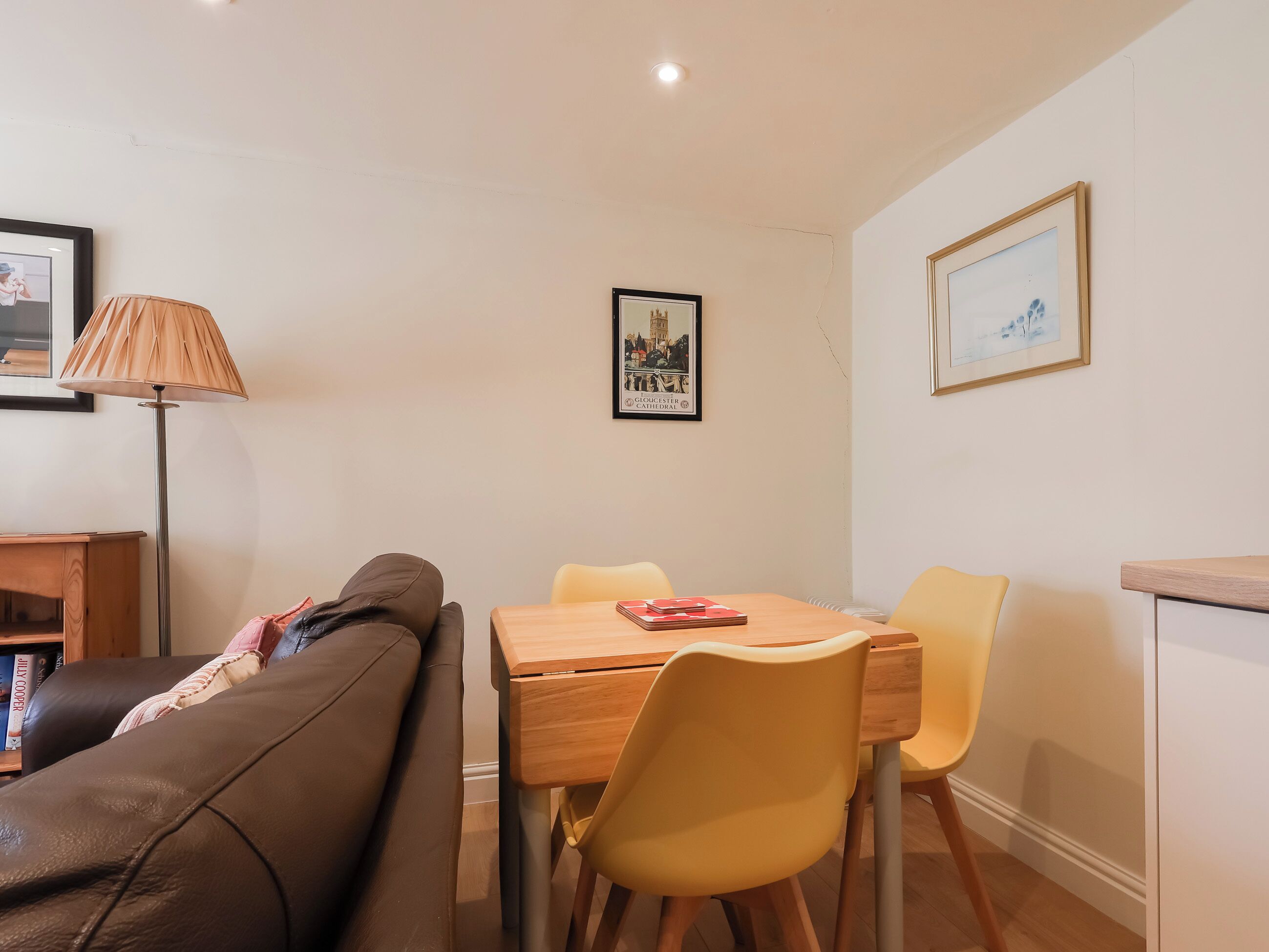 Een woonkamer met een bruine leren bank, een houten eettafel met gele stoelen, een lamp en lijstwerken aan de muur.