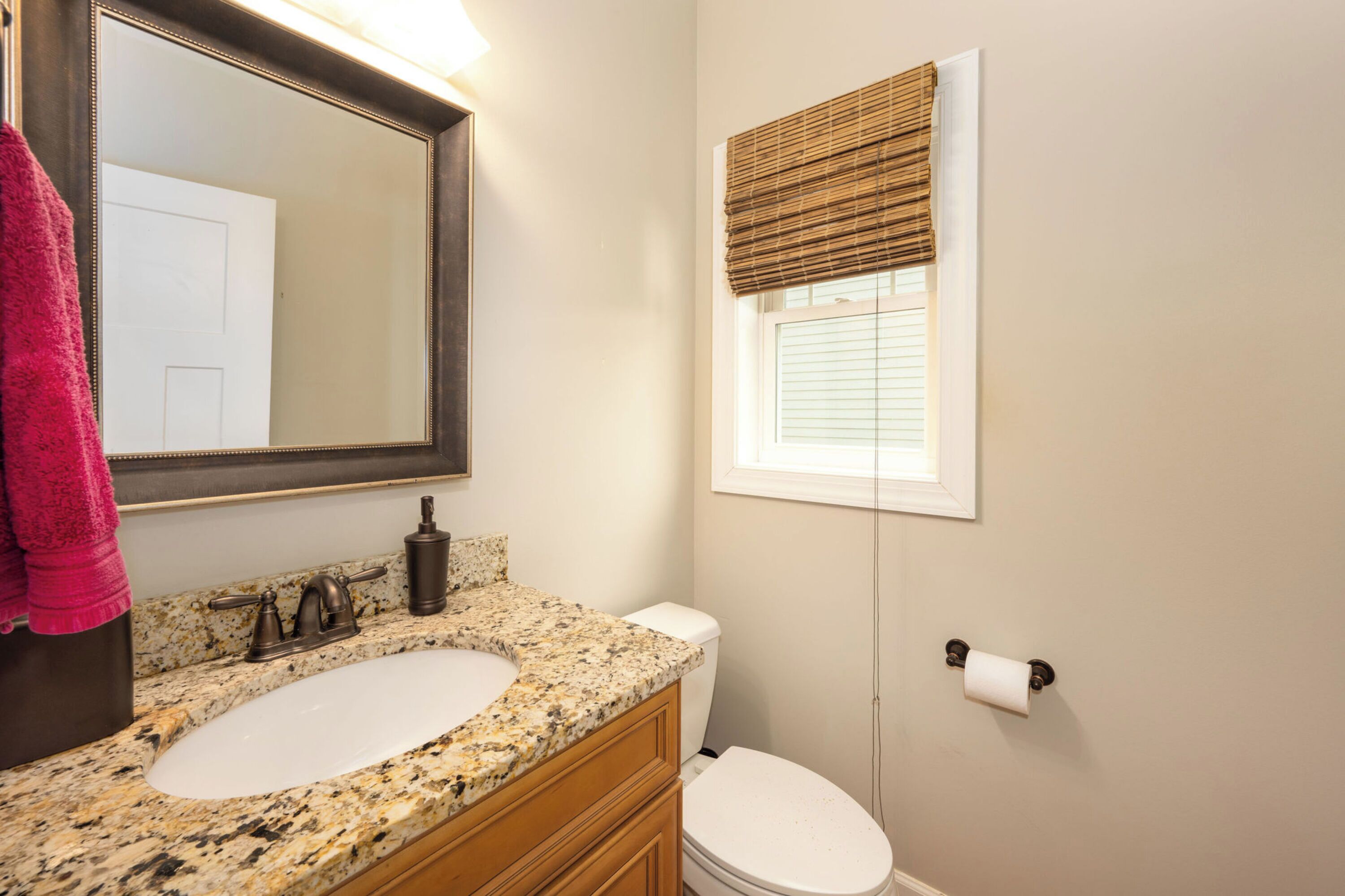 A bathroom with a granite sink, a mirror, a toilet, and a window with blinds.