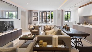 A modern living room with a sofa, coffee table, and a TV. There is a kitchen area with a dining table and bar stools.