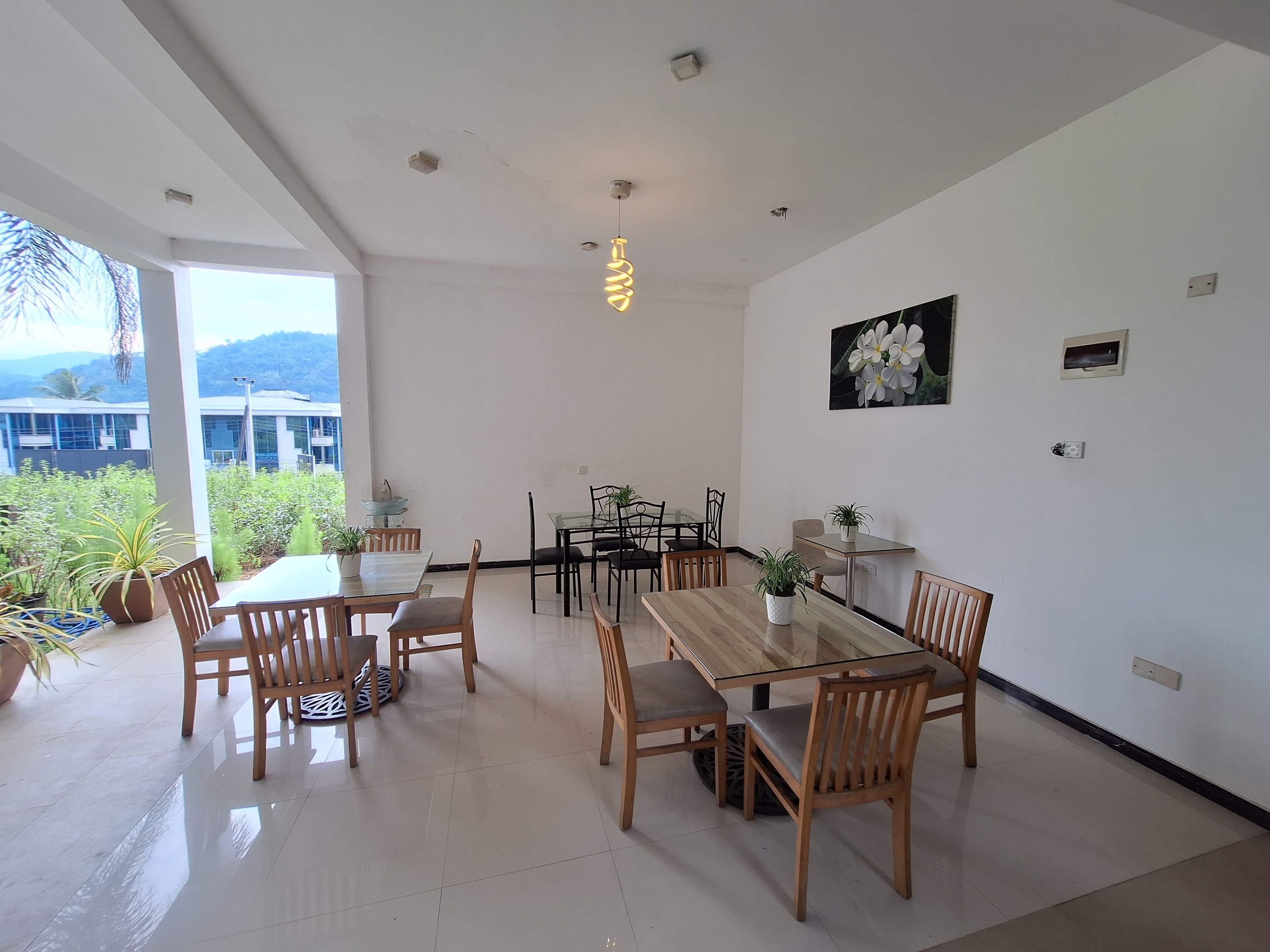 A spacious dining area with a glass table and wooden chairs, surrounded by white walls and a large window offering a view of greenery and a building.