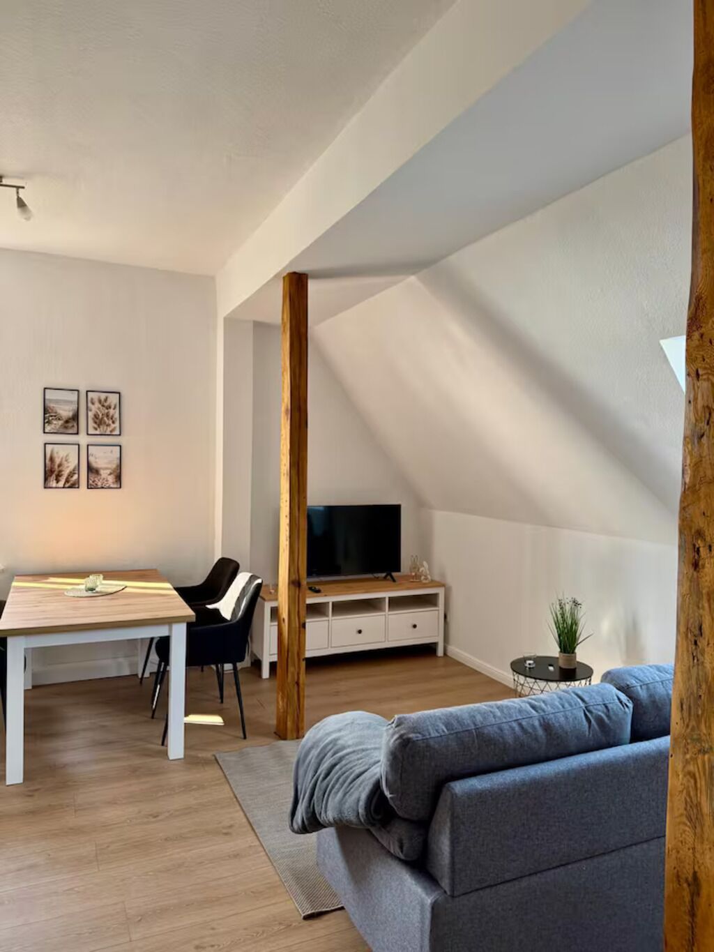 A modern loft apartment with a dining area, a gray sofa, wooden beams, and a TV in the background.