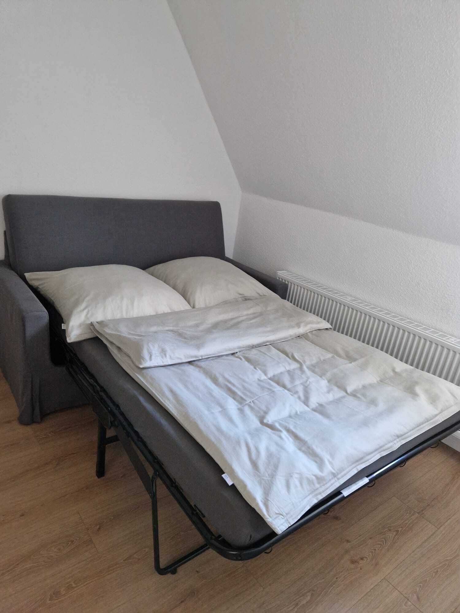 Un lit compact dans une pièce dotée d’un plafond en pente, d’un radiateur et d’un canapé gris.