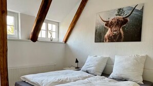 Un dormitorio con una cama, paredes blancas, una ventana y un cuadro de una vaca de las Tierras Altas.
