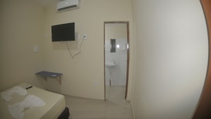 Ein kleines Hotelzimmer mit einem Bett, einem an der Wand befestigten Fernseher, einem Schreibtisch und einem durchgegangenen Tür sichtbaren Badezimmer.