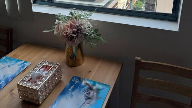 A room with a large window overlooking a cityscape, a wooden table with a vase of flowers, and a decorative box.