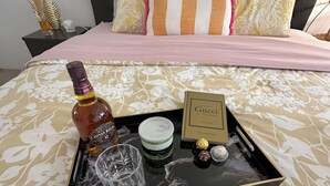 A neatly arranged tray on a bed with a floral bedspread, containing a bottle of Chivas Regal, a glass, a jar of Nutella, and a box of Gucci chocolates.