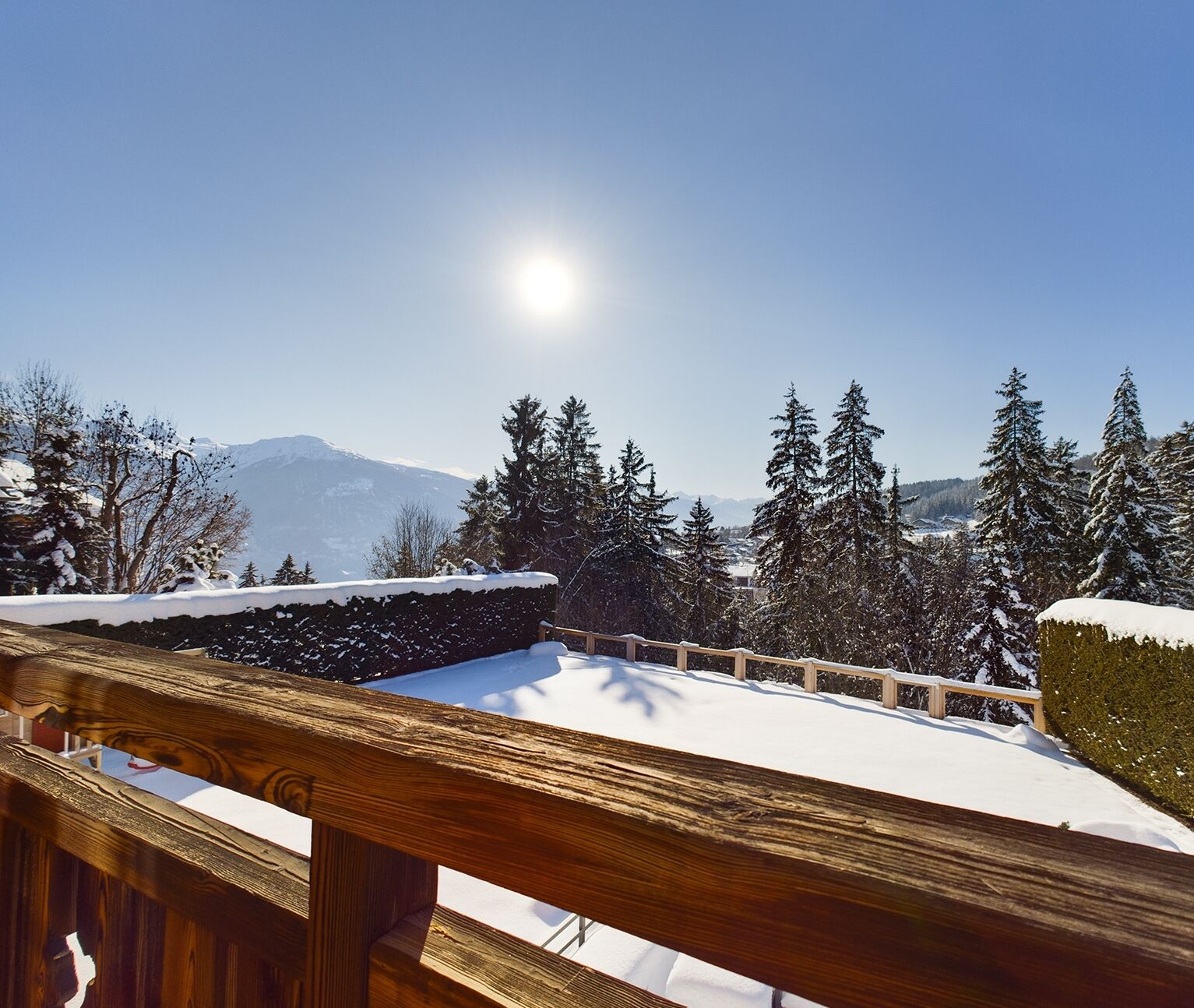 Une terrasse en bois, le sol couvert de neige et des arbres à feuilles persistantes en arrière-plan.
