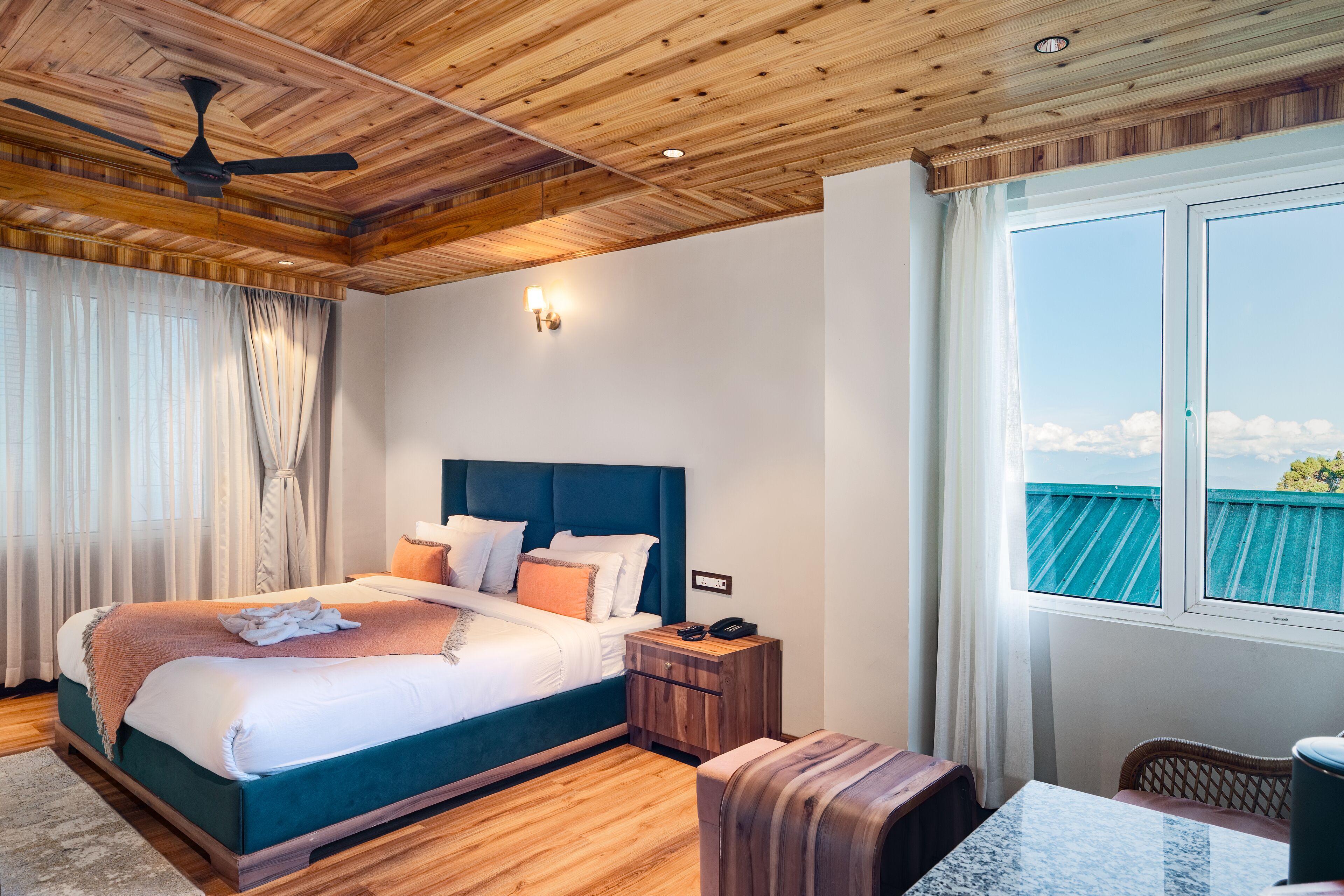 Un dormitorio con una cama grande, techo de madera, una mesita de noche, una mesa y vista al cielo a través de una ventana.