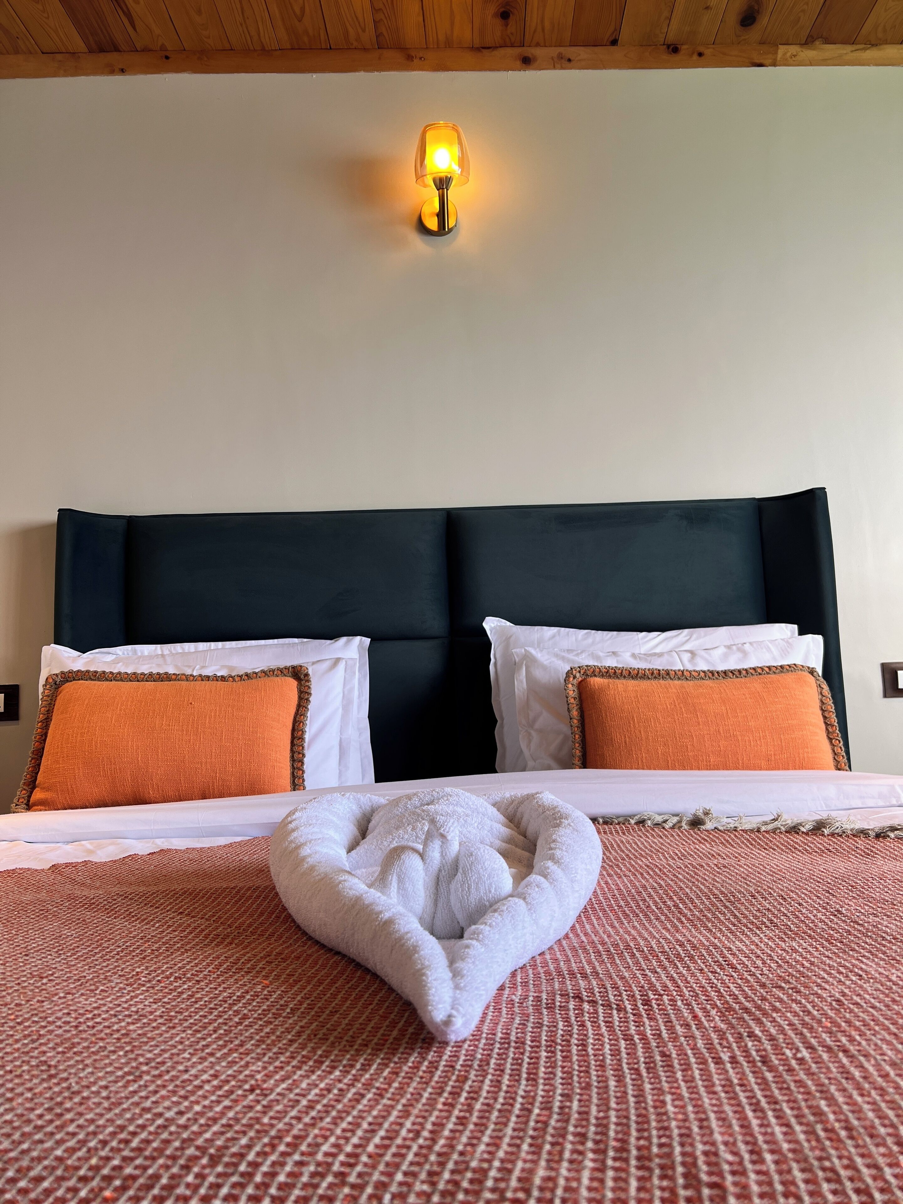 A neatly made bed with a white and orange blanket, two white pillows, and an orange throw pillow. A white towel is placed on the bed.