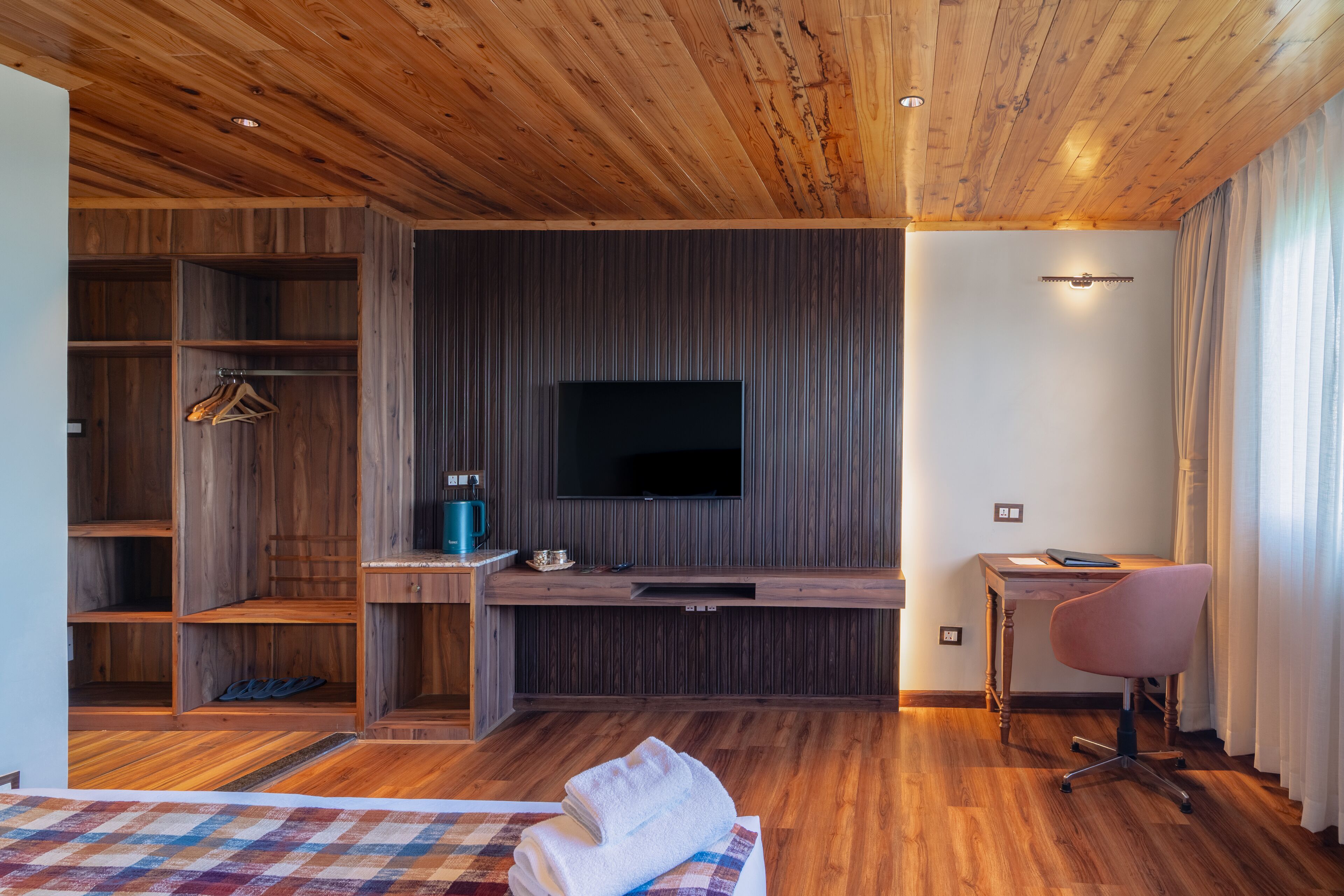Habitación de hotel moderna con suelo de madera, televisor de pantalla plana montado en una pared oscura, escritorio con silla y armario con espacio para colgar.