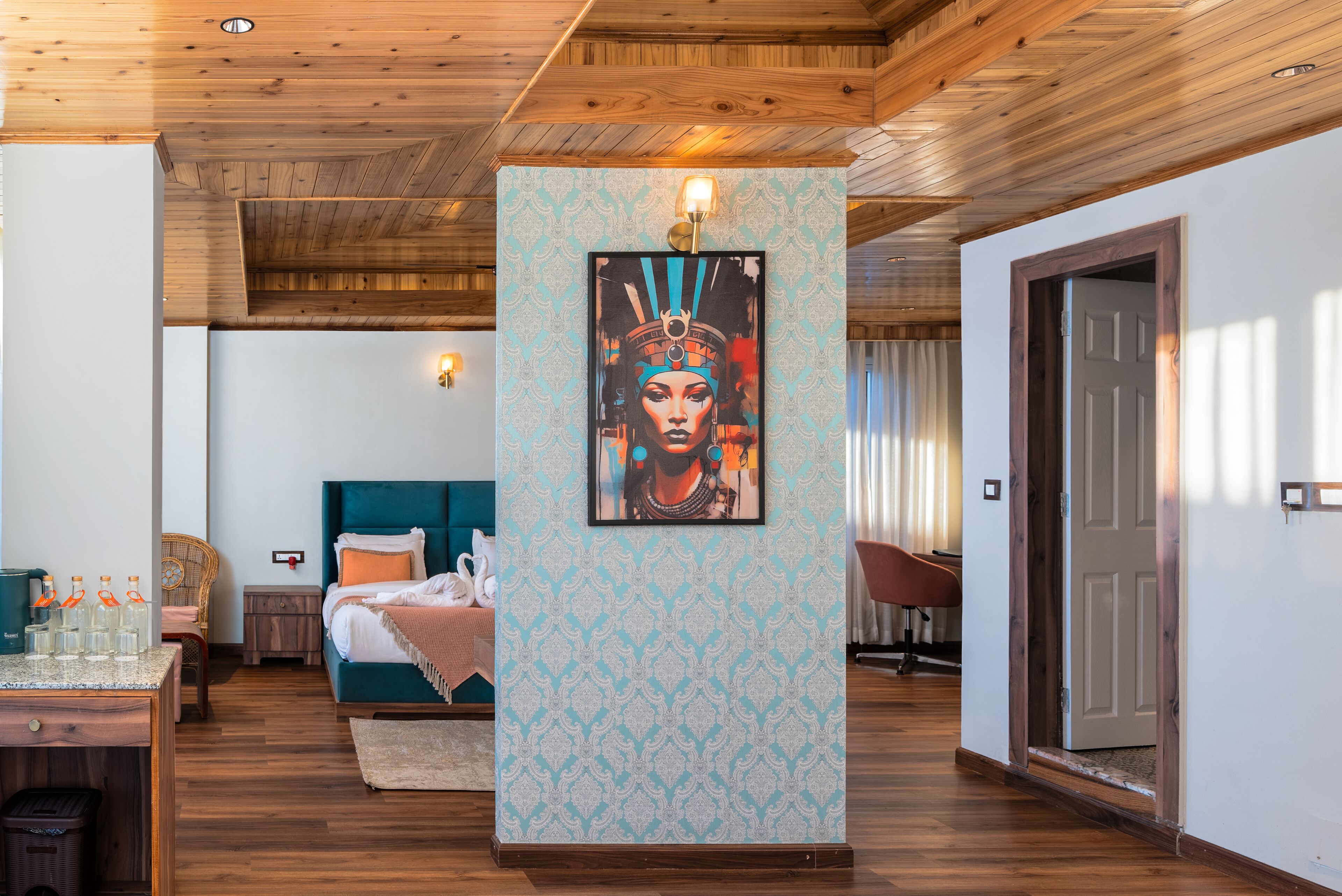 Habitación de hotel con techo de madera, cama con ropa de cama color turquesa, una obra de arte colgada en la pared, una mesita con botellas y una silla de mimbre.