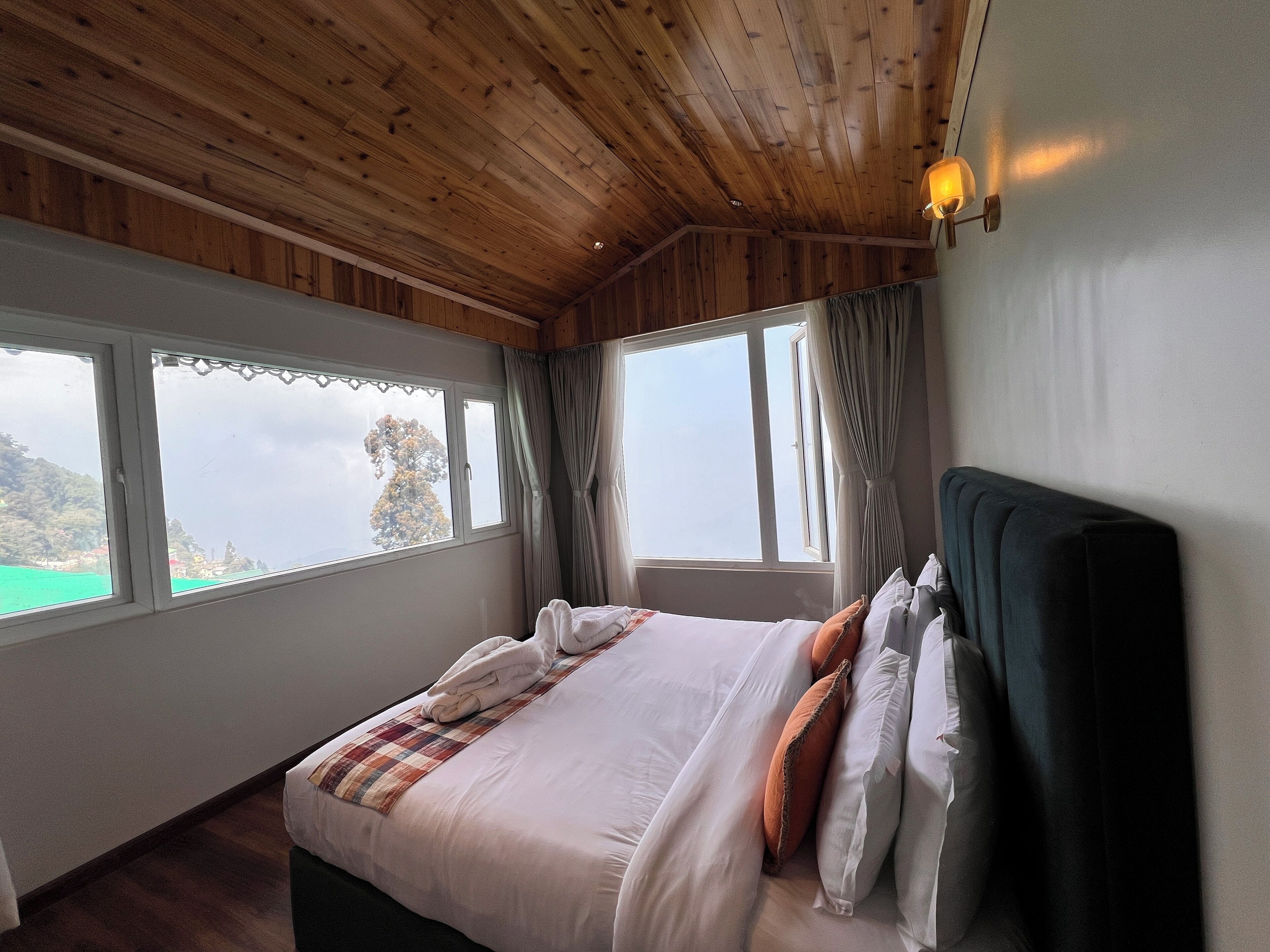 A bedroom with a wooden ceiling, a bed with white bedding and orange pillows, two windows with curtains, and a view of greenery outside.