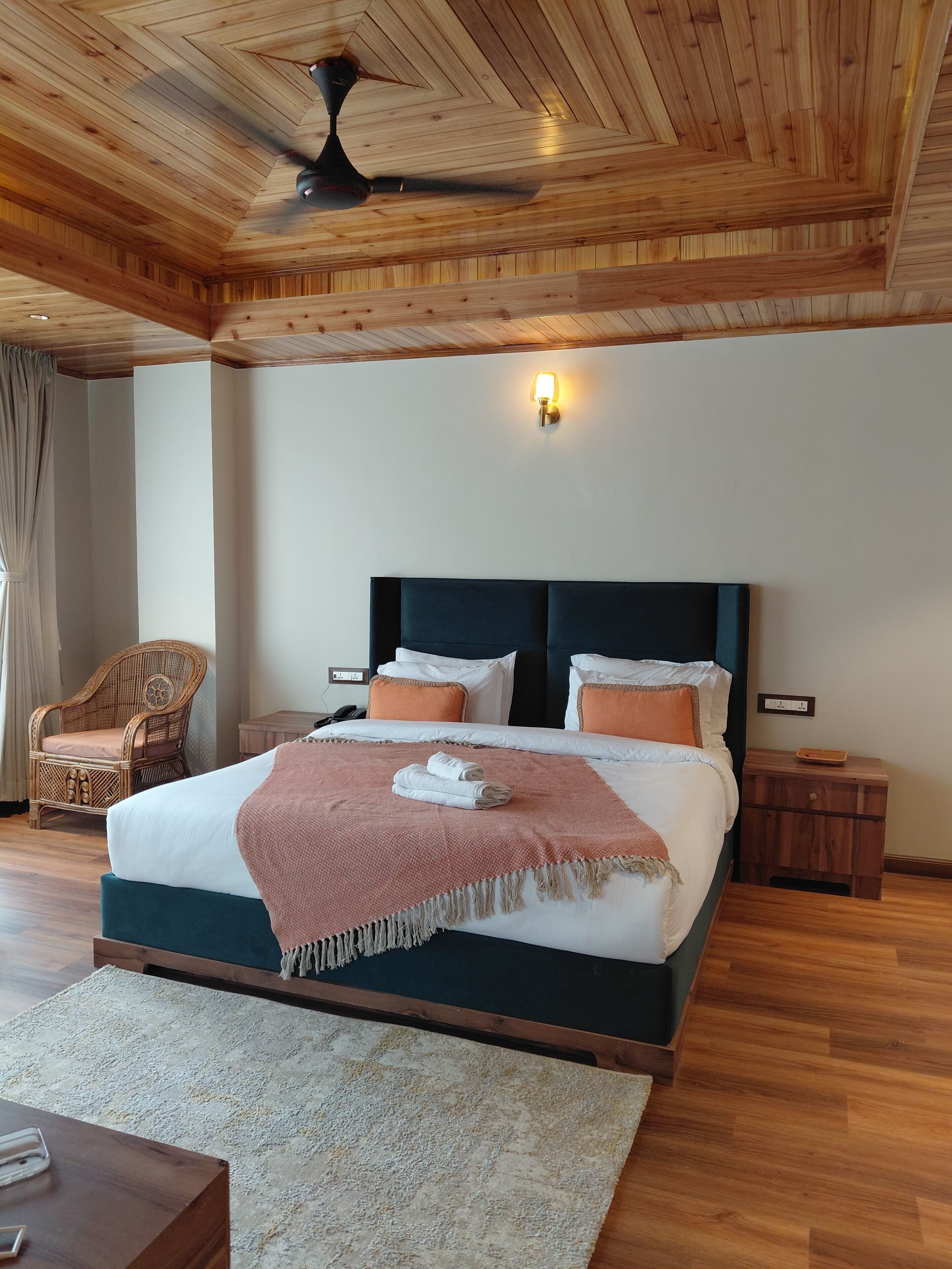 A spacious bedroom with a large bed, a wooden ceiling, a wall-mounted light, a wicker chair, and a bedside table.