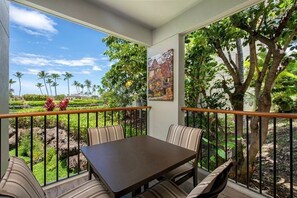 A balcony with a table and chairs, a view of a garden, and a painting on the wall. - 1BR-Beautiful Ground Floor, Ocean View, Mauna Lani Resort (Kamuela)