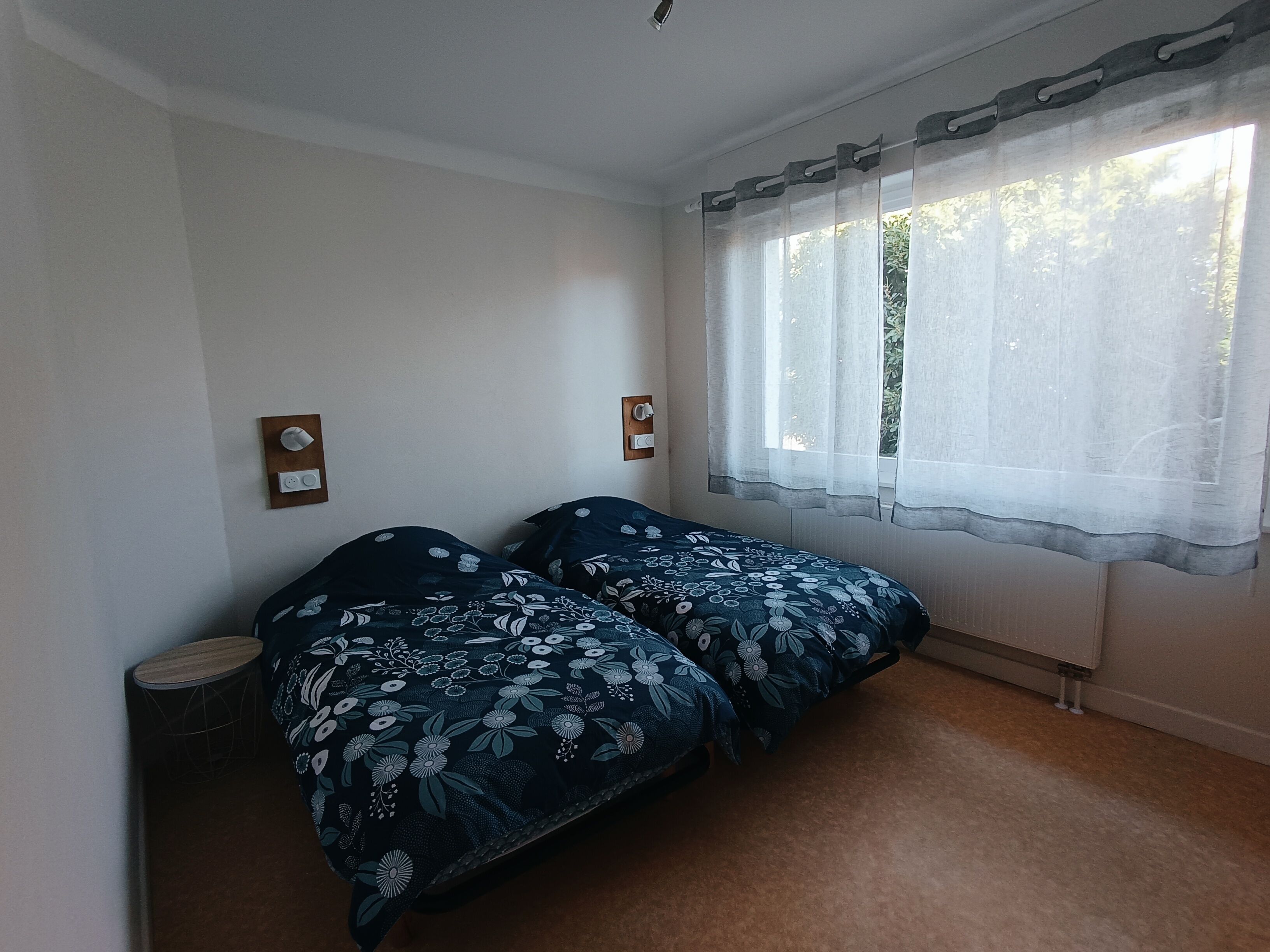 A bedroom with two beds, a window with sheer curtains, and a wall-mounted light switch.