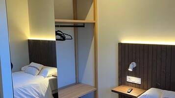 Habitación de hotel compacta con dos camas, armario empotrado, mesita de noche con lámpara y aire acondicionado.