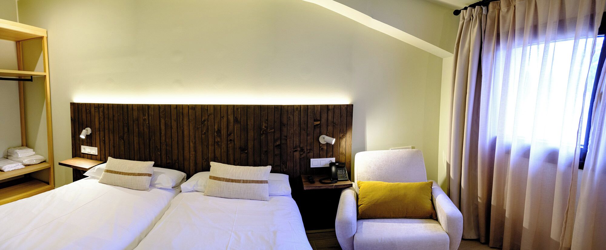 Habitación de hotel con dos camas, cabecera de madera, mesita de noche con lámpara, una silla con almohada amarilla y una ventana con cortinas.