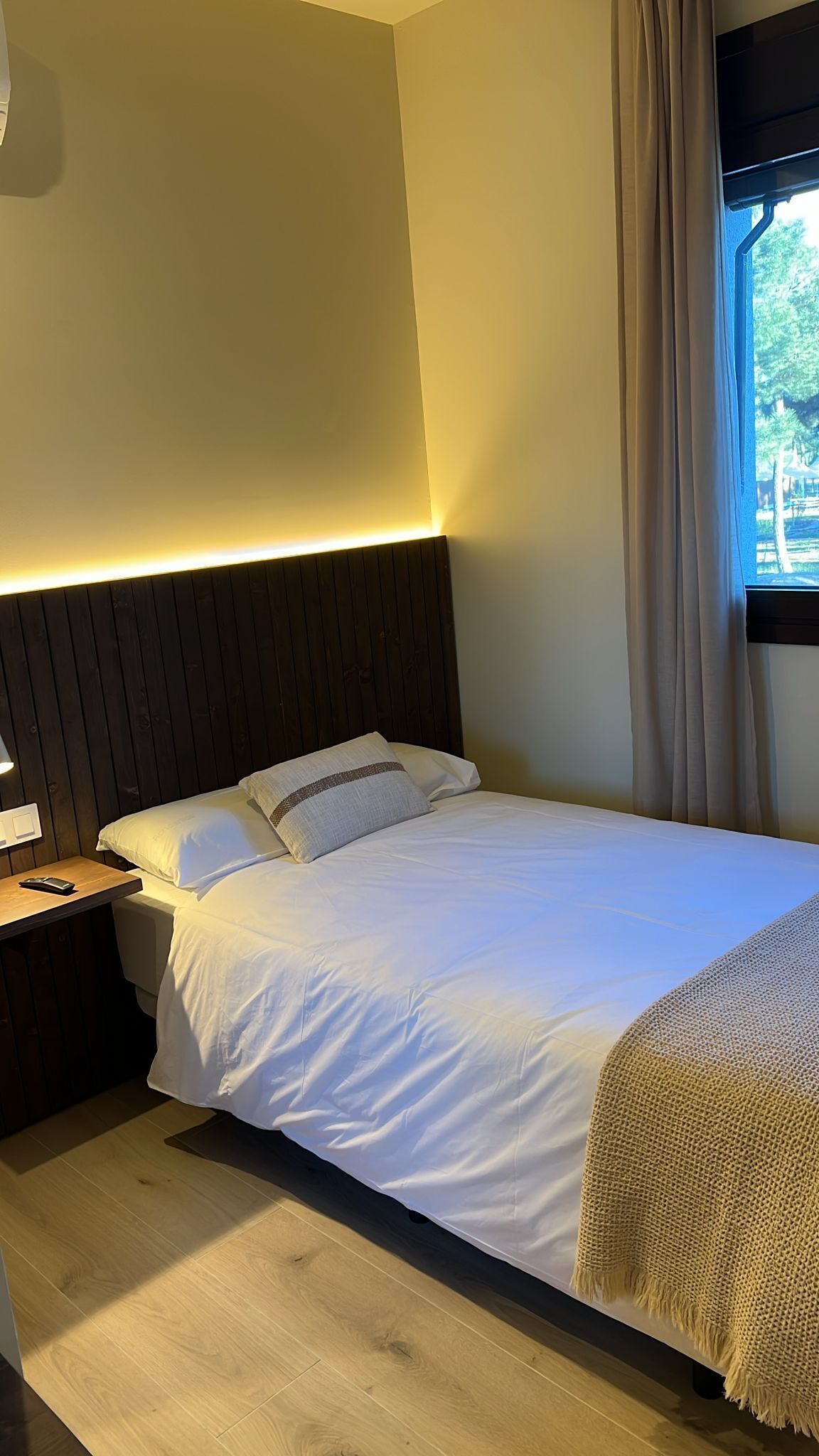 A neatly made bed with white linens and a dark headboard, a bedside table with a lamp, and a window with curtains.