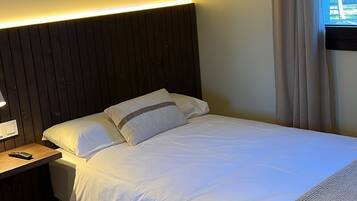A neatly made bed with white linens and a dark headboard, a bedside table with a lamp, and a window with curtains.
