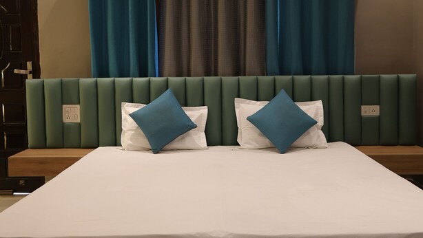 A modern hotel room with a large bed, green upholstered headboard, and blue curtains. - River View Resort (Haridwar)