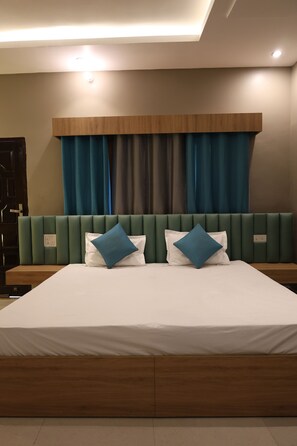A modern hotel room with a large bed, green upholstered headboard, and blue curtains. - River View Resort (Haridwar)