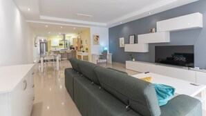 En moderne stue med en grå sofa, et fladskærms-tv og et hvidt tv-møbel. I baggrunden ses et køkken med hvide skabe og en gul væg.