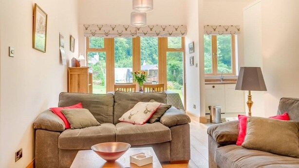 A cozy living room with a sofa set, a coffee table, and a lamp. Large windows offer a view of the outdoors.