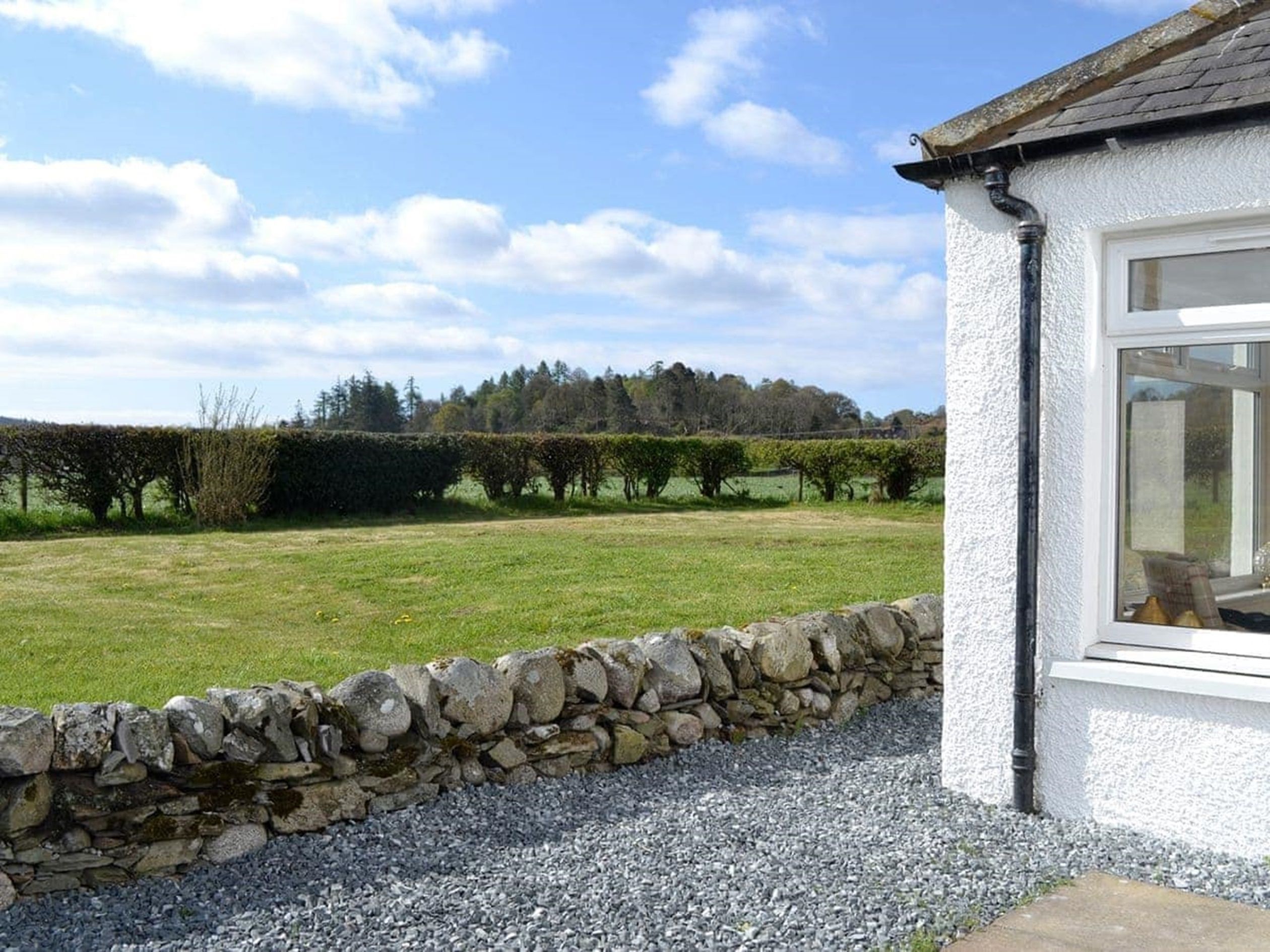 A stone wall, a gravel path, and a grassy field with a hedge in the background.